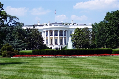[ai] The White House, a large white neoclassical building, is seen from a grassy area with manicured lawns and colorful flower beds in the foreground, under a clear blue sky with a few clouds.